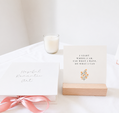 Affirmation card displayed in a wooden holder beside a Hopeful Romantic Art gift box with a pink ribbon and candle