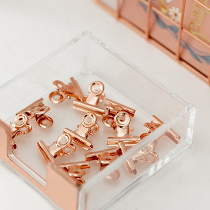 Rose gold bulldog clips in a clear acrylic tray on a bright stationery desk